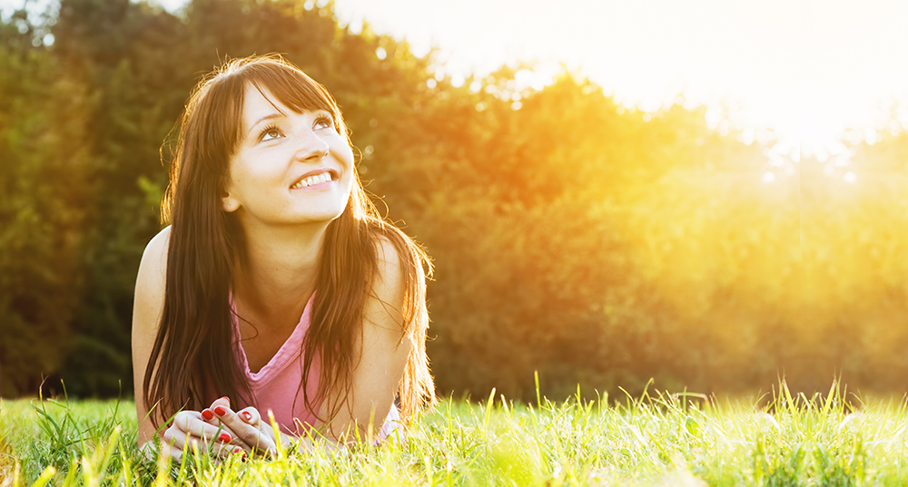 Young beautiful woman smiling and lying on the grass at summer sunset looking at the sky. Natural happiness, fun and harmony.